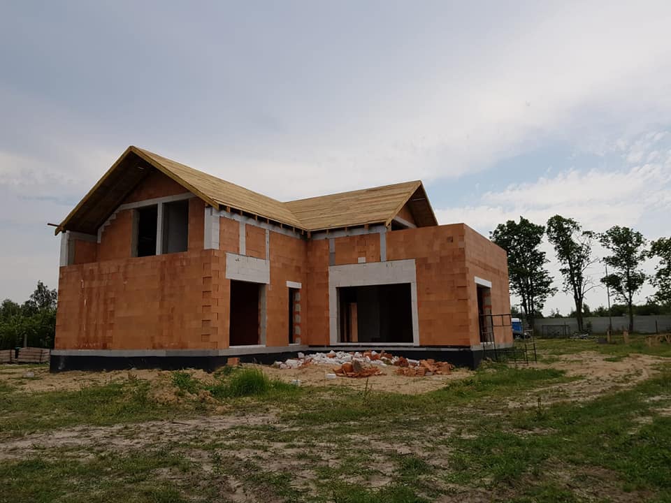 Surowy budynek jednorodzinny w trakcie budowy: ściany z czerwonej cegły, drewniana konstrukcja dachu, widoczne otwory okienne i drzwiowe, teren budowy z gruzem i piaskiem.