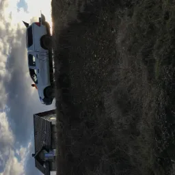 Biały samochód terenowy z podniesionym zawieszeniem i flagą na dachu, zaparkowany na nierównym terenie w pobliżu domu jednorodzinnego z ciemną dachówką, na tle zachmurzonego nieba.