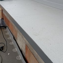 EKOOBUDOWA SPÓŁKA Z OGRANICZONĄ ODPOWIEDZIALNOŚCIĄ - Szare obróbki blacharskie na dachu z widocznym narzędziem do zgrzewania i fragmentem muru z cegły.