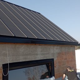 EKOOBUDOWA SPÓŁKA Z OGRANICZONĄ ODPOWIEDZIALNOŚCIĄ - Nowoczesny dach z blachy na surowym budynku, dekarz w trakcie prac, widoczne narzędzia i materiały przygotowane do montażu. Okno z czarną ramą odbija drzewa.