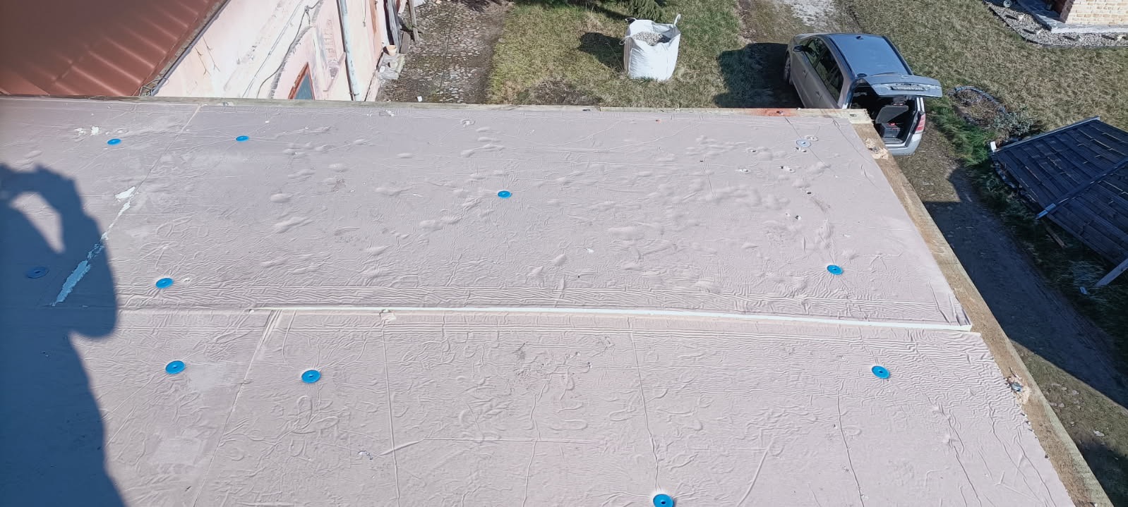 Widok z góry na płaski dach pokryty jasną membraną dachową, z widocznymi niebieskimi elementami mocującymi, zarys cienia osoby w lewym dolnym rogu, w tle samochód kombi z otwartym bagażnikiem...