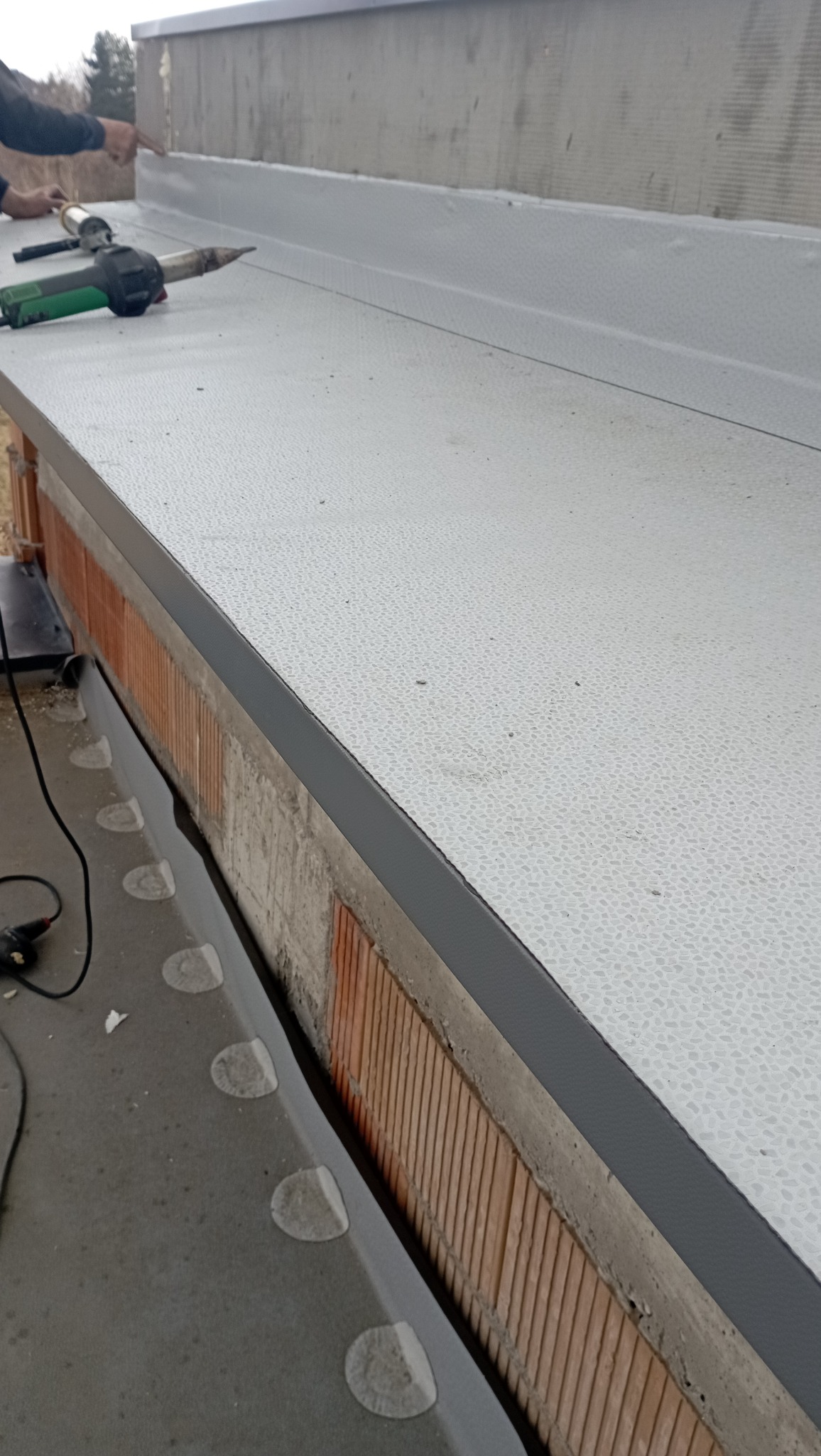 Szare obróbki blacharskie na dachu z widocznym narzędziem do zgrzewania i fragmentem muru z cegły.