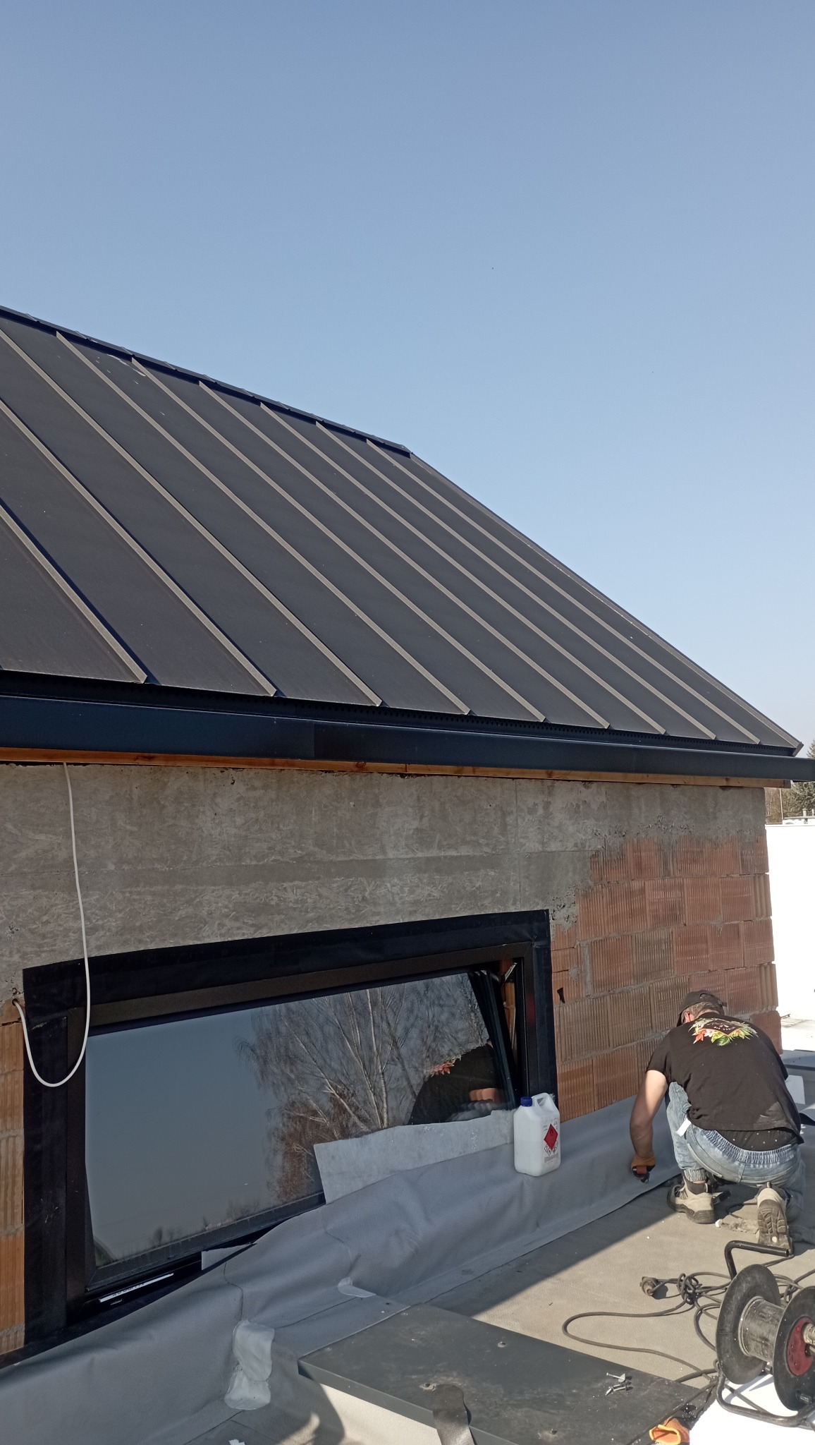 Nowoczesny dach z blachy na surowym budynku, dekarz w trakcie prac, widoczne narzędzia i materiały przygotowane do montażu. Okno z czarną ramą odbija drzewa.