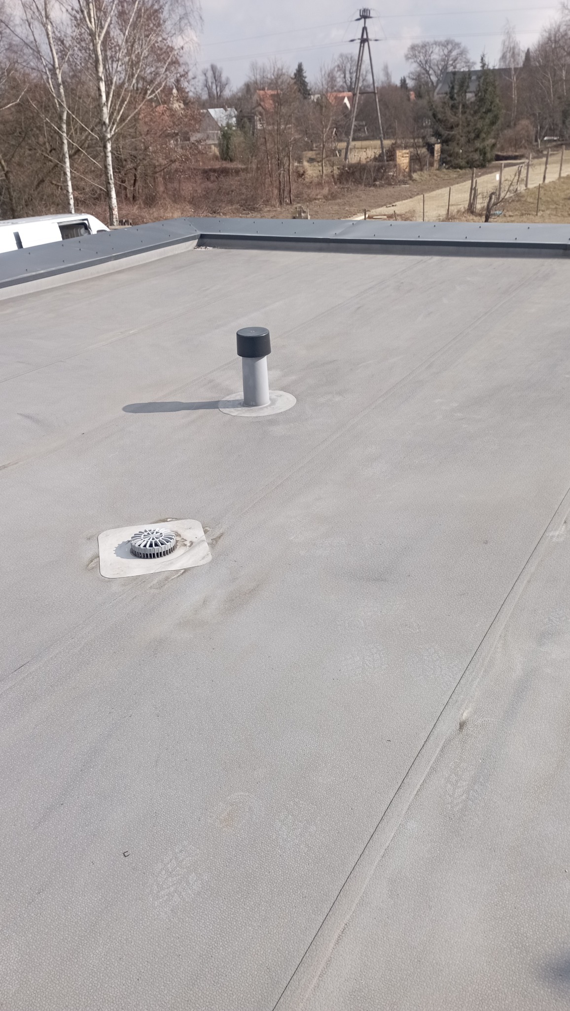 Szary, płaski dach pokryty papą termozgrzewalną z dwoma elementami: kominkiem wentylacyjnym z czarną nasadką i wpustem dachowym z osadnikiem liści.