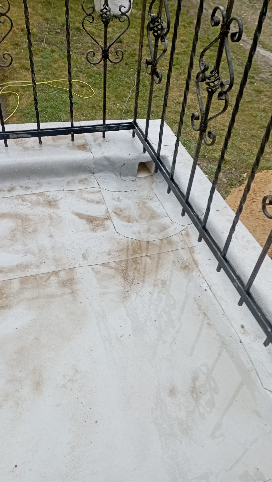 Biała, popękana powierzchnia balkonu z widoczną rynną odpływową i ozdobną, metalową balustradą z kutymi elementami w kształcie serc.