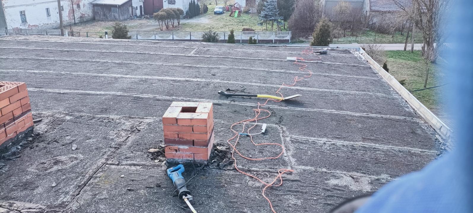 Demontaż starego komina z cegły na pokrytym papą dachu, widoczna piła szablasta, łopata i przewód elektryczny, w tle zabudowania.