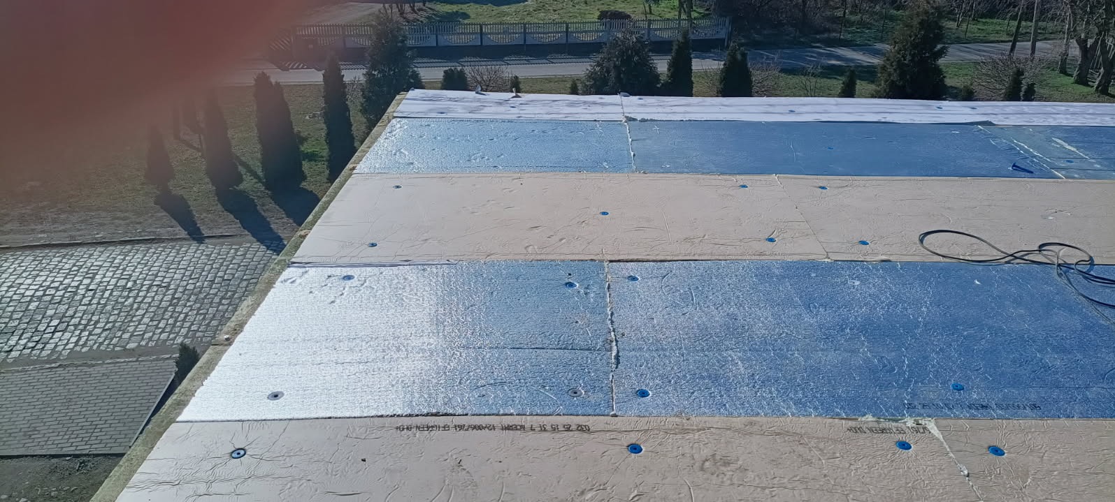 Widok z góry na warstwy izolacyjne z folią aluminiową i jasnymi płytami na dachu budynku, z widocznymi niebieskimi mocowaniami i kablem elektrycznym, w tle zieleń i drzewa.