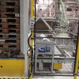 DAN-PRO-AUTOMATIC Sylwester Dana - Żółta barierka ochronna z siatki zabezpiecza robota przemysłowego, widoczne palety w tle, element sterujący z przyciskami i lampką kontrolną.