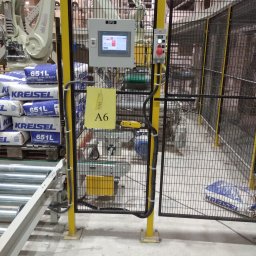 DAN-PRO-AUTOMATIC Sylwester Dana - Zabezpieczona klatka z żółtymi słupkami i czarną siatką otaczająca zautomatyzowaną linię produkcyjną z robotem przemysłowym; widoczne worki z zaprawą gipsową Kreisel 651L i panel sterowania SPS.