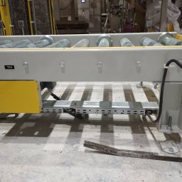 DAN-PRO-AUTOMATIC Sylwester Dana - Fragment automatycznej linii produkcyjnej z rolkowym podajnikiem, widoczne elementy konstrukcyjne, napęd elektryczny w żółtej obudowie oraz przewody w peszlu.