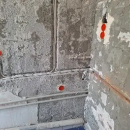 Ściana w trakcie remontu z widocznymi przewodami elektrycznymi, puszkami instalacyjnymi zabezpieczonymi pomarańczowymi dekielkami, gniazdkiem elektrycznym oraz rurami. Ściana surowa, z widocznymi...