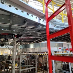 Skomplikowana konstrukcja metalowa z widocznymi elementami automatyki przemysłowej, korytami kablowymi i fragmentem czerwonej drabiny, prawdopodobnie w hali magazynowej.