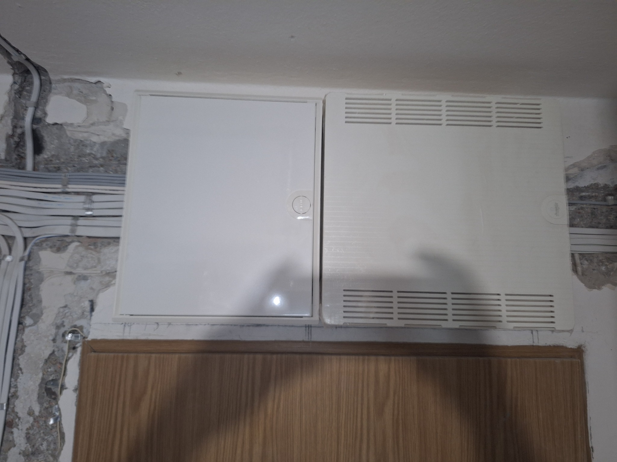 Dwa białe panele dostępowe, jeden gładki z okrągłym zamkiem, drugi wentylowany, zamontowane w ścianie nad drewnianą boazerią, widoczne kable elektryczne w listwach na ścianie z ubytkami tynku.