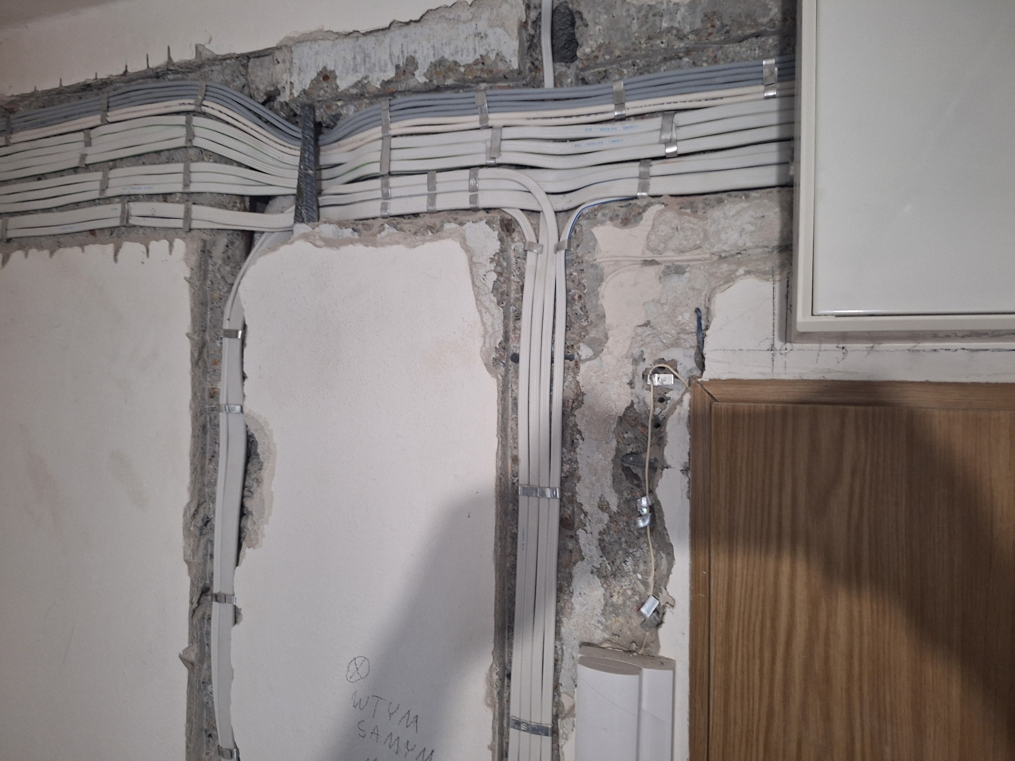 Ściana w trakcie remontu z widocznymi przewodami elektrycznymi ułożonymi w korytach i przymocowanymi metalowymi uchwytami, obok widoczny fragment drewnianych drzwi.