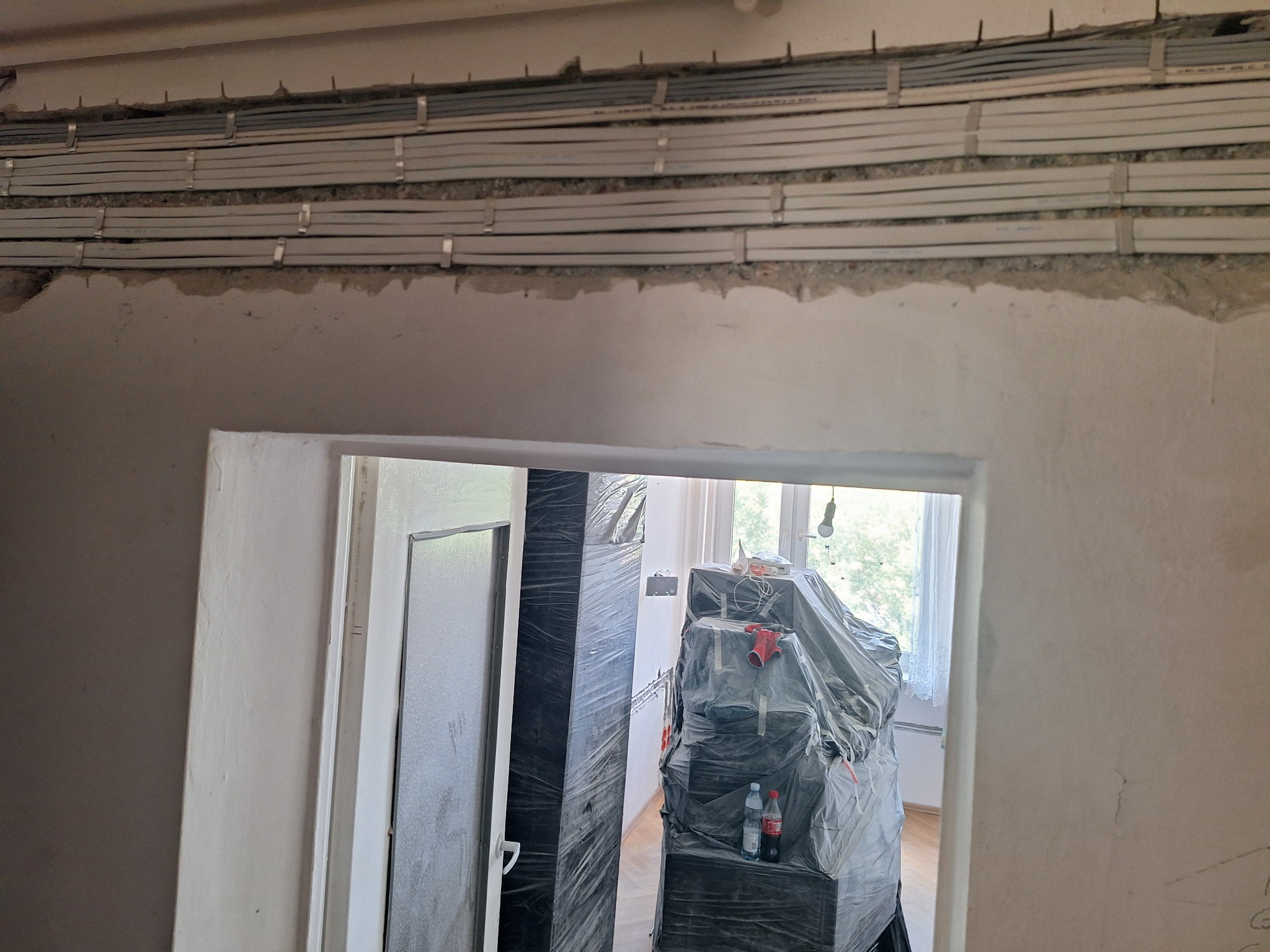 Rzędy szarych przewodów elektrycznych przymocowanych do ściany za pomocą metalowych uchwytów, widoczne przez otwór w ścianie w trakcie remontu, z zakrytymi meblami w tle.