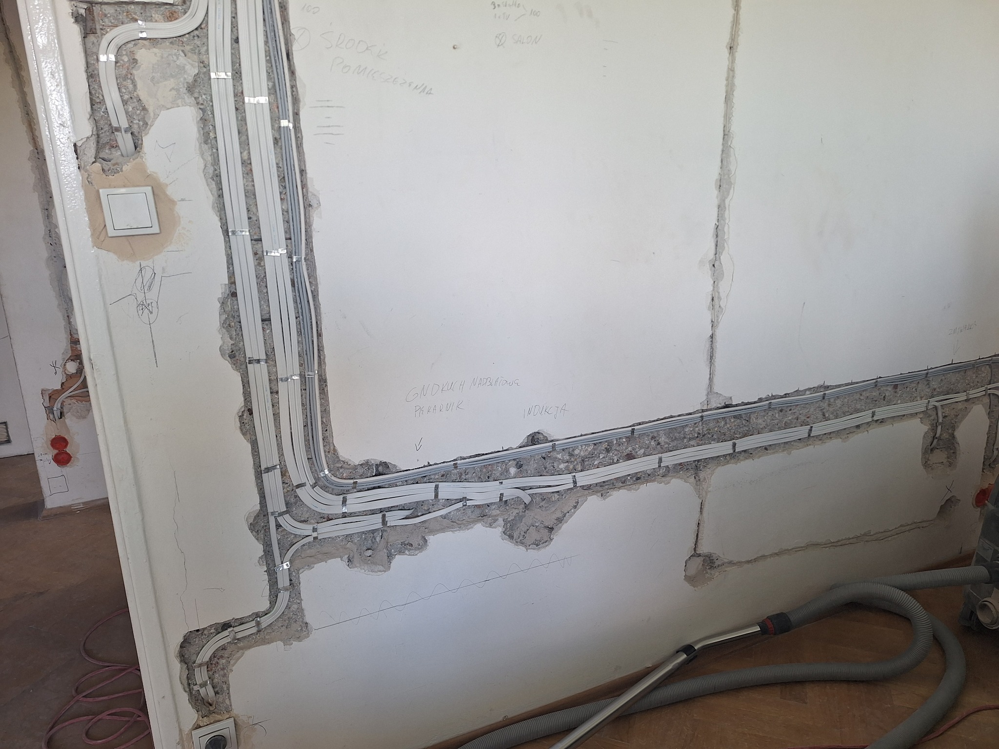 Wnętrze w trakcie remontu: widoczne kable elektryczne prowadzone w bruzdach w ścianie, zabezpieczone metalowymi uchwytami, zarysowane na ścianie notatki i podłączenie odkurzacza przemysłowego.