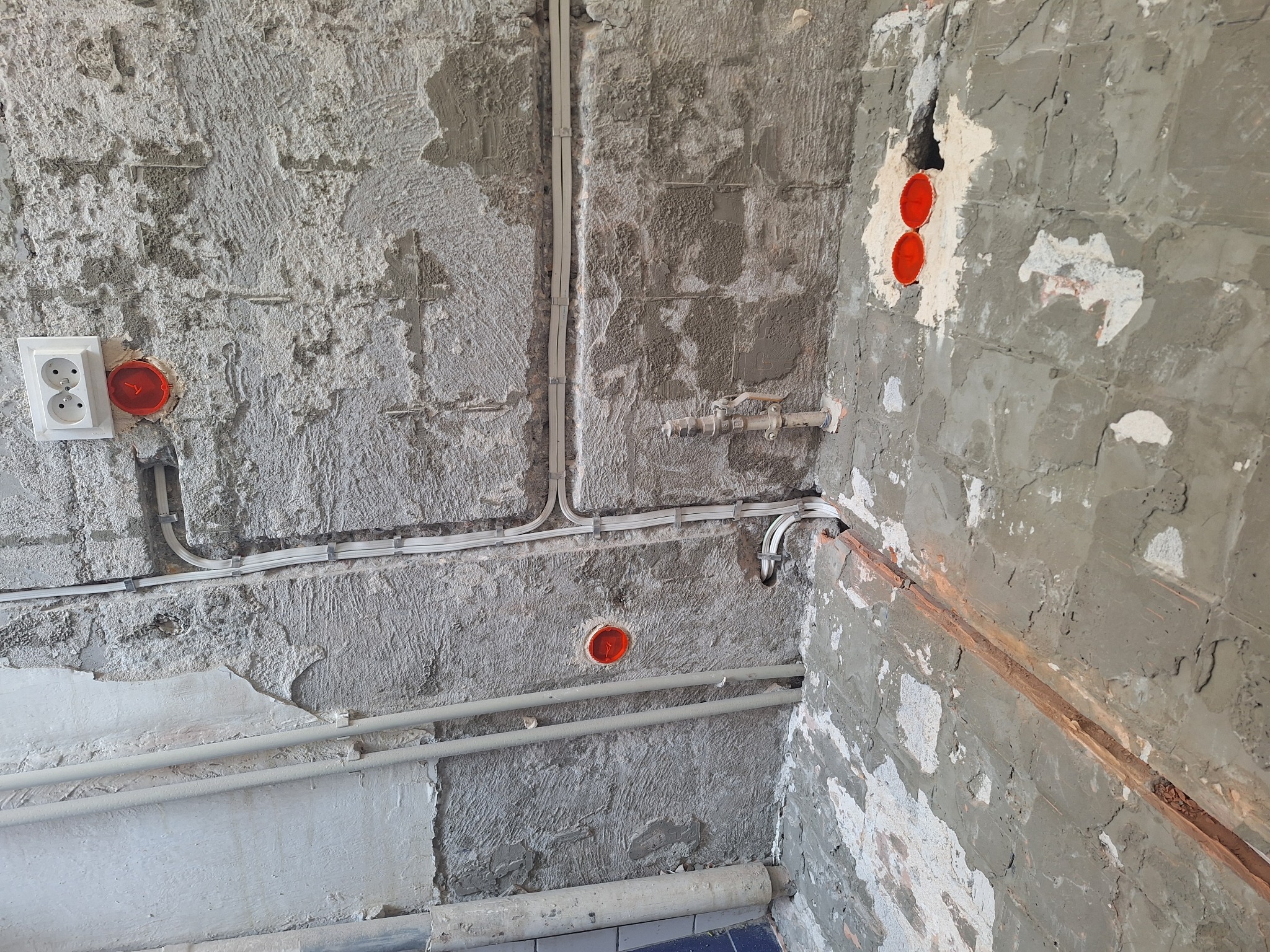 Ściana w trakcie remontu z widocznymi przewodami elektrycznymi, puszkami instalacyjnymi zabezpieczonymi pomarańczowymi dekielkami, gniazdkiem elektrycznym oraz rurami. Ściana surowa, z widocznymi...