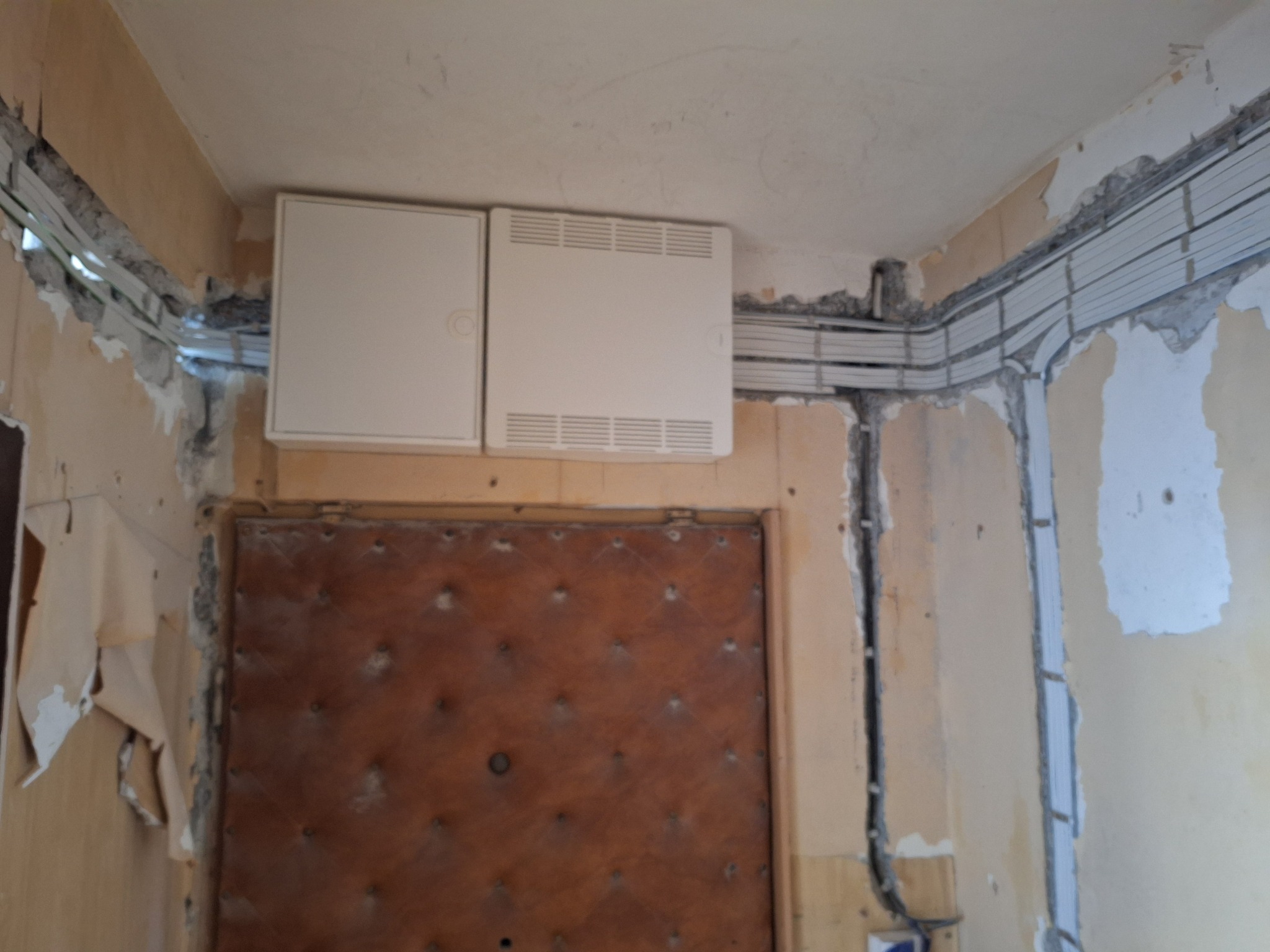 Dwa białe panele elektryczne zamontowane nad brązowymi drzwiami w trakcie remontu, widoczne kable w korytkach przy suficie i ściany z resztkami tapet.