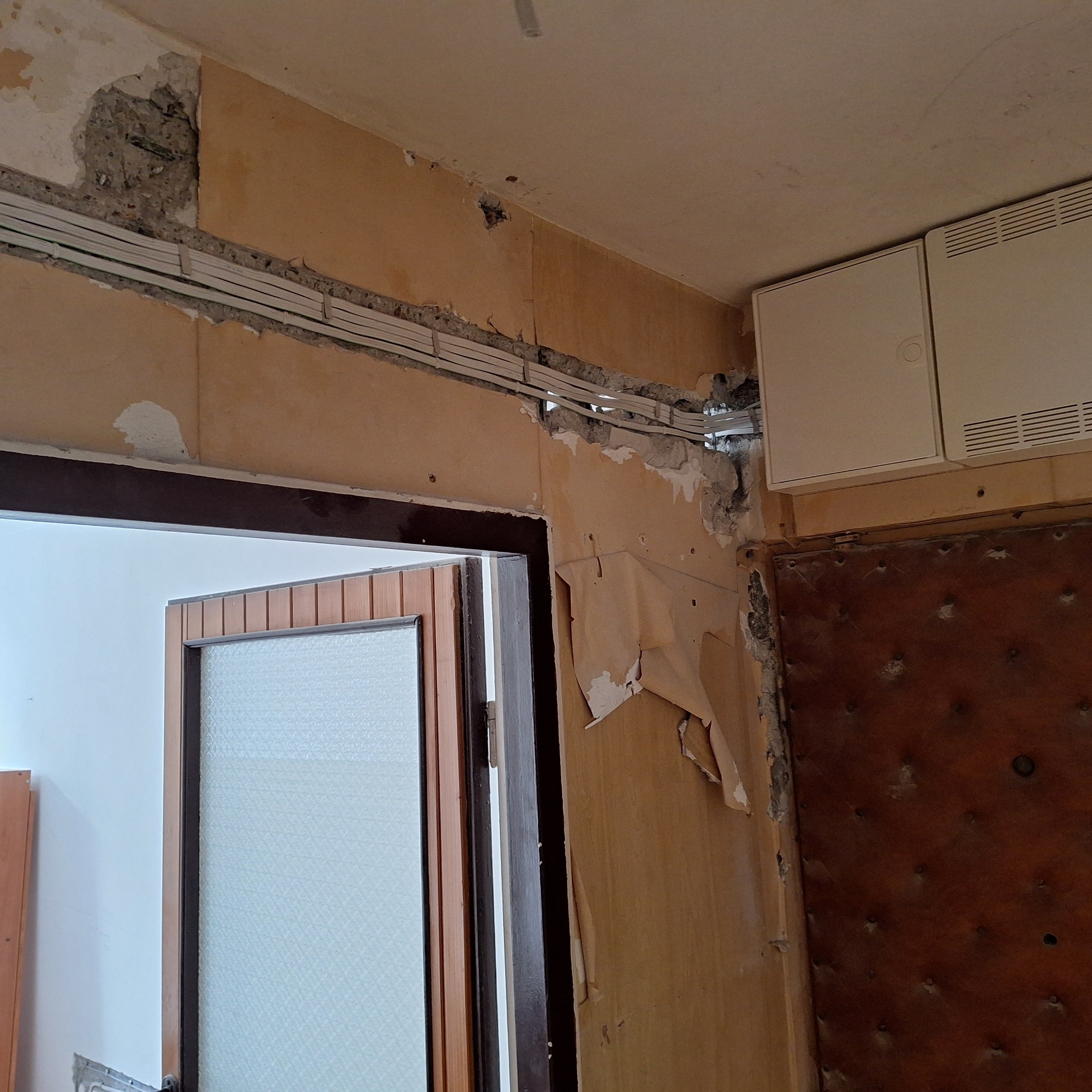 Kable elektryczne ułożone w bruździe ściennej nad futryną drzwi, ściana w trakcie remontu z odchodzącą tapetą i widoczną skrzynką elektryczną.