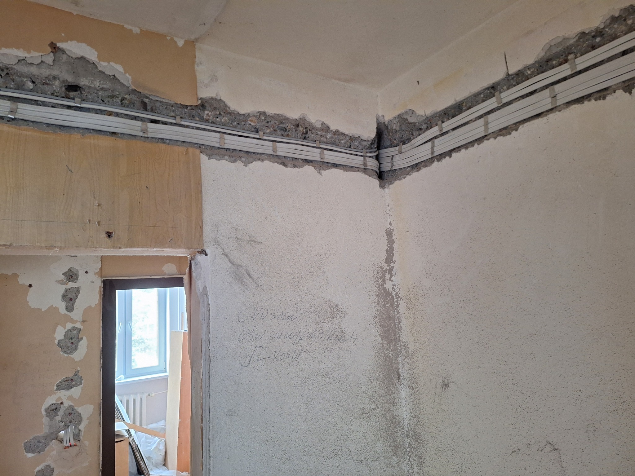 Wnętrze w trakcie remontu, widoczne kable elektryczne w korytach zamontowane wzdłuż ściany z surowym wykończeniem, częściowo usunięta tapeta, notatki ołówkiem na ścianie, drzwi do innego...