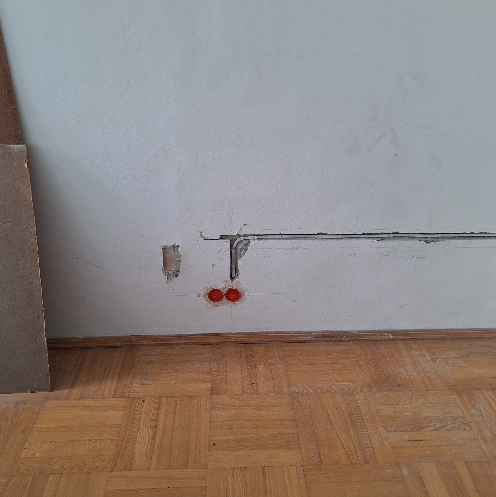 Biała ściana z wyżłobieniami pod przewody elektryczne i dwoma czerwonymi puszkami instalacyjnymi, widoczny fragment drewnianej podłogi parkietowej i płyta z boku.