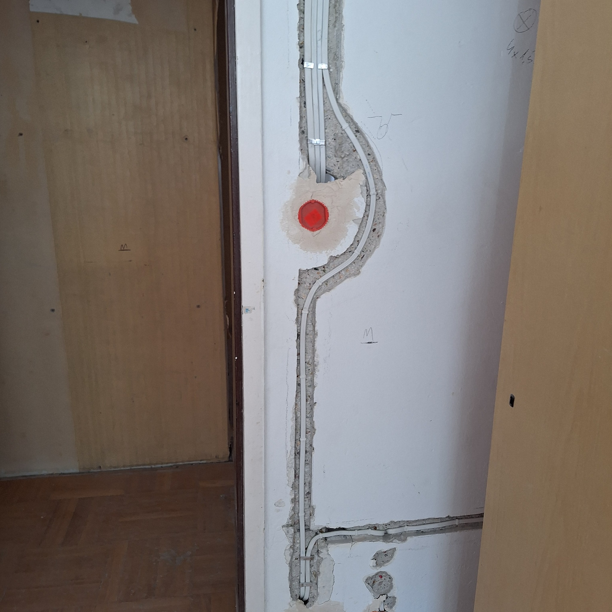 Białe kable elektryczne prowadzone w bruzdach w ścianie, zabezpieczone metalowymi uchwytami, z widoczną czerwoną puszką instalacyjną i śladami po kuciu w tynku.