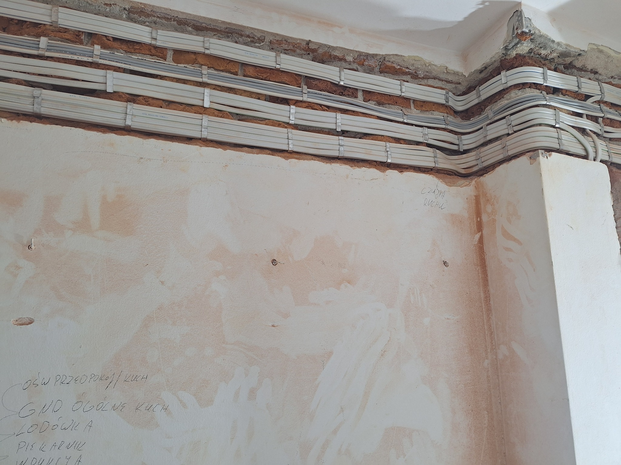 Kable elektryczne ułożone w korytkach na ścianie z odsłoniętą cegłą, widoczne napisy ołówkiem na ścianie opisujące obwody.