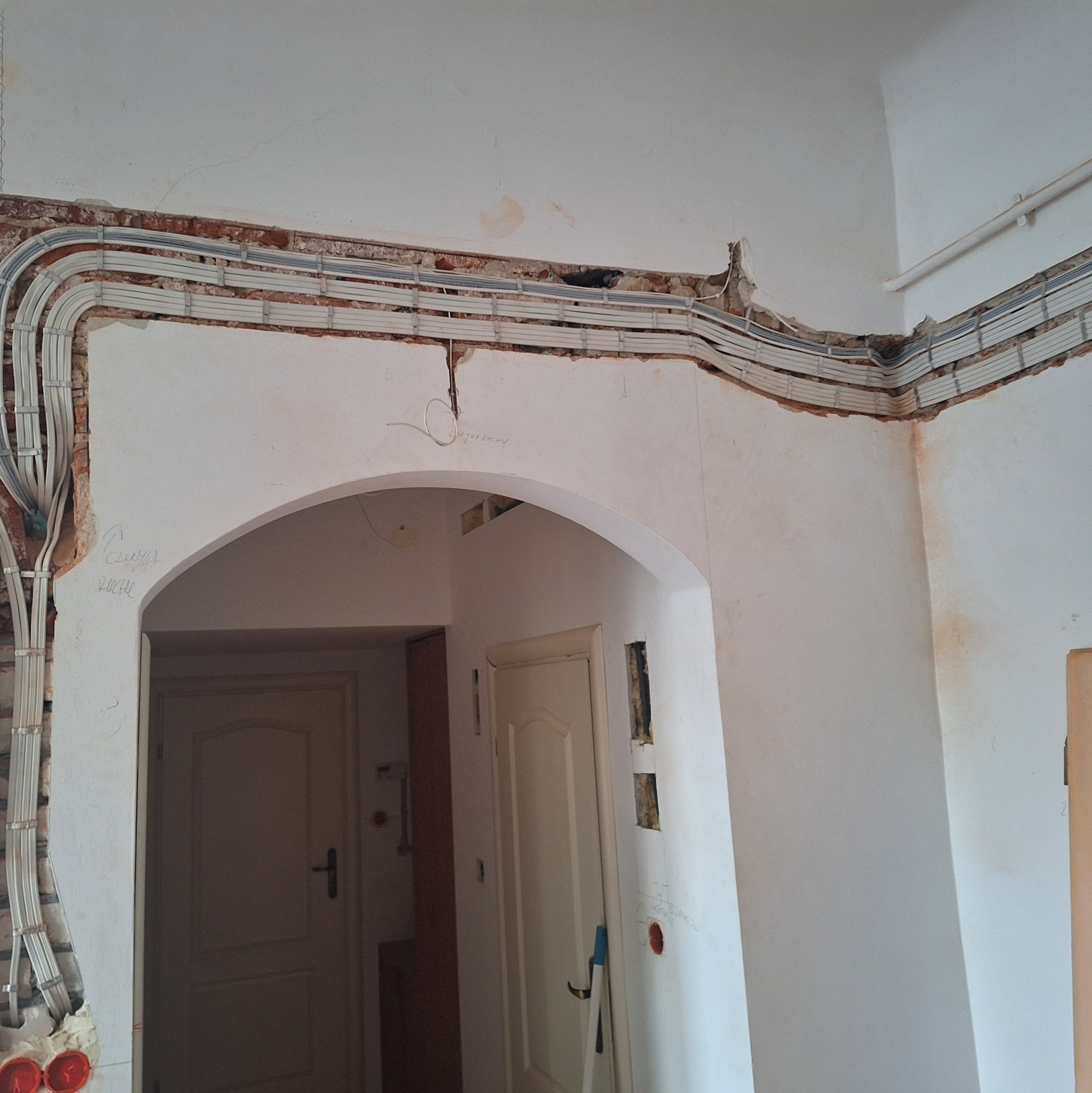Montaż przewodów elektrycznych w bruzdach pod sufitem, widoczne kable w szarych rurkach, ściany w trakcie remontu, widoczne fragmenty cegieł i niedomalowane powierzchnie, przejście łukowe do innego...