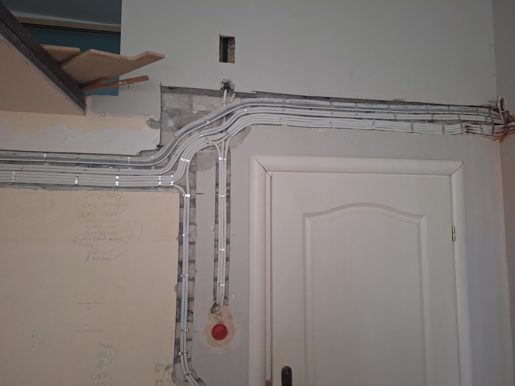 Białe kable elektryczne w peszlach ułożone wzdłuż ściany nad drzwiami, widoczne notatki ołówkiem na ścianie i niedokończone wykończenie ściany.