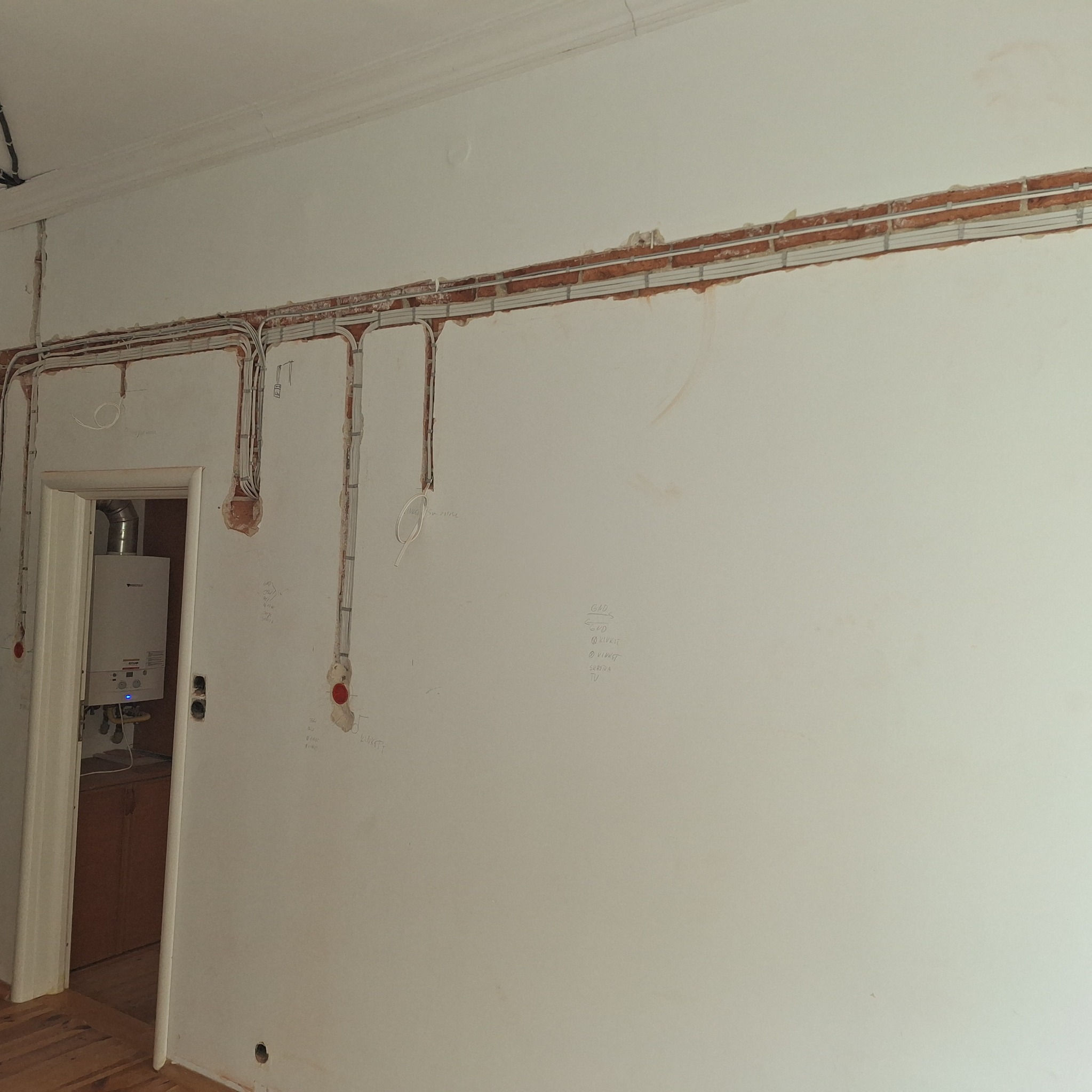 Ściana w trakcie kładzenia instalacji elektrycznej w Serocku, widoczne kable w korytkach, ślady po kuciu w ścianie i piec gazowy w tle.