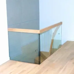 Szklana balustrada z drewnianą poręczą zabezpieczająca schody, widok z góry na jasną podłogę.