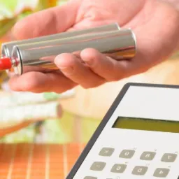 Dwie metalowe elektrody podłączone czerwonymi przewodami do urządzenia diagnostycznego trzymane w dłoniach, na tle jasnego wnętrza z kwiatami.