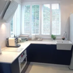 Jasna kuchnia z granatowymi szafkami pod blatem, białymi żaluzjami w oknach i drewnianą podłogą w odcieniu bieli.