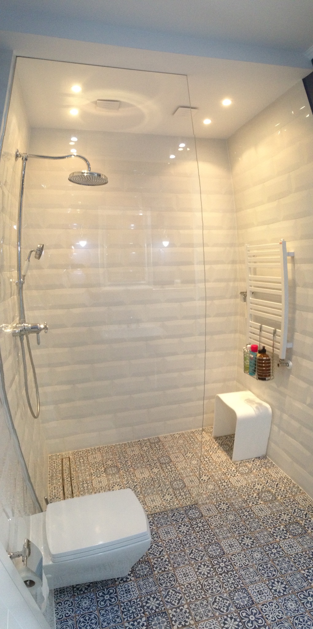 Nowoczesna kabina prysznicowa walk-in z deszczownicą, białą ceramiką i dekoracyjną, wzorzystą podłogą w odcieniach szarości i beżu.