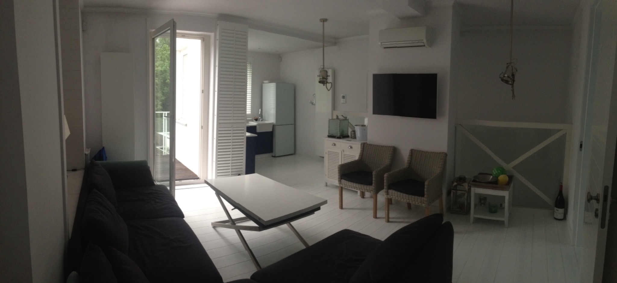 Jasne wnętrze apartamentu z widokiem na balkon, ciemnoszara sofa, biały stolik kawowy z regulowaną wysokością, rattanowe krzesła i aneks kuchenny w tle.