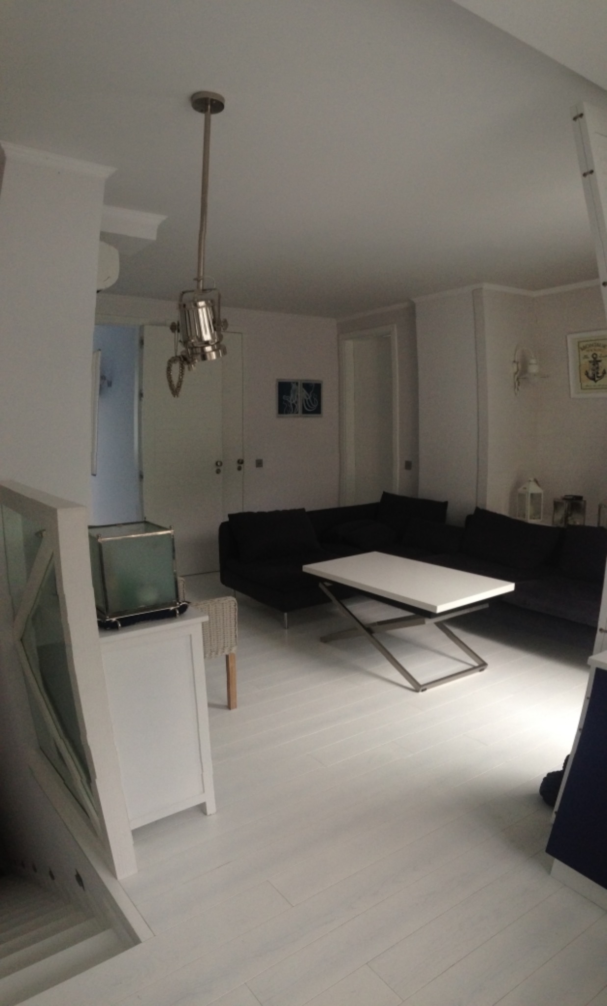 Jasne wnętrze apartamentu nadmorskiego z ciemną sofą narożną, białym stolikiem kawowym na metalowej podstawie i dekoracyjną lampą wiszącą w stylu marynistycznym.