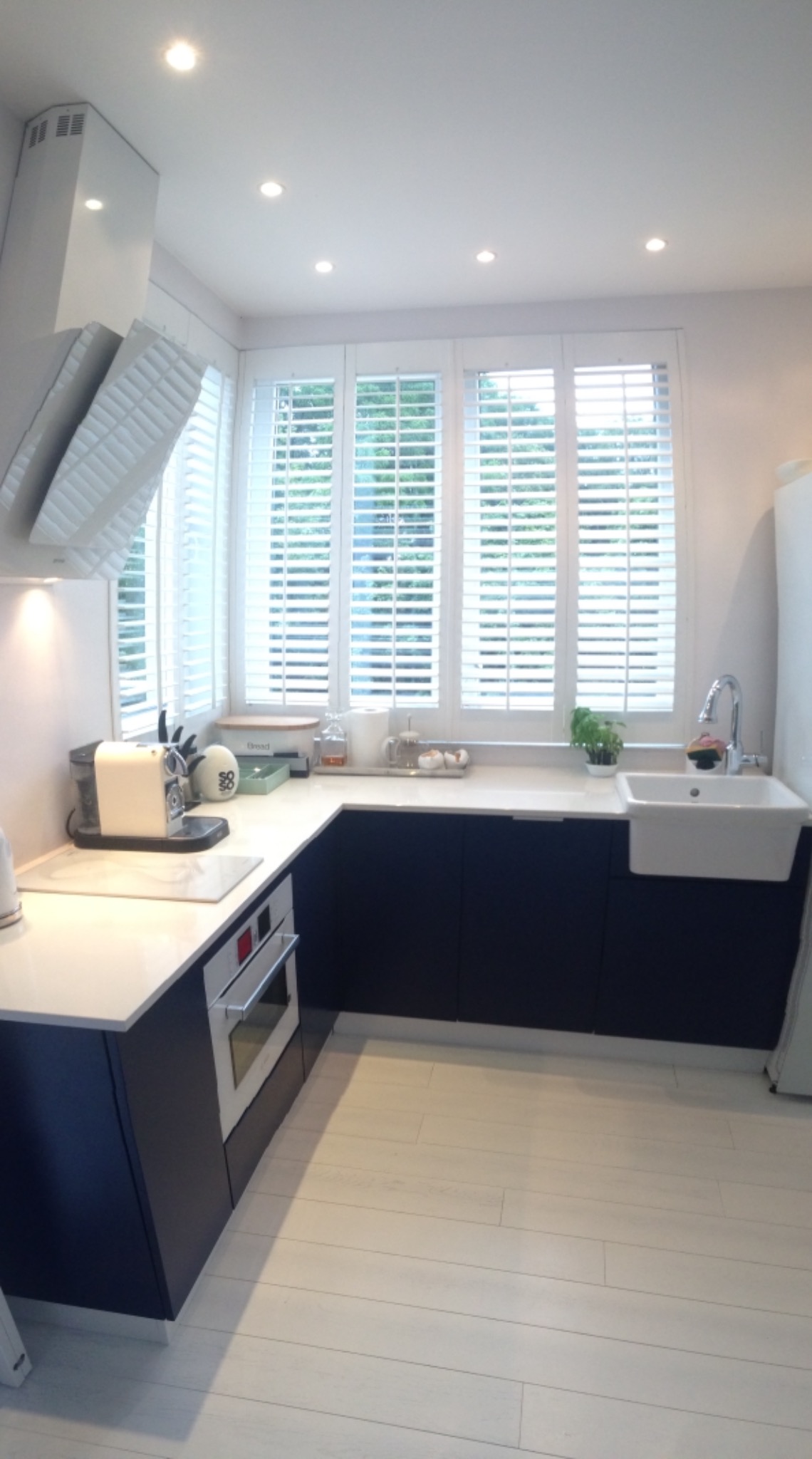 Jasna kuchnia z granatowymi szafkami pod blatem, białymi żaluzjami w oknach i drewnianą podłogą w odcieniu bieli.
