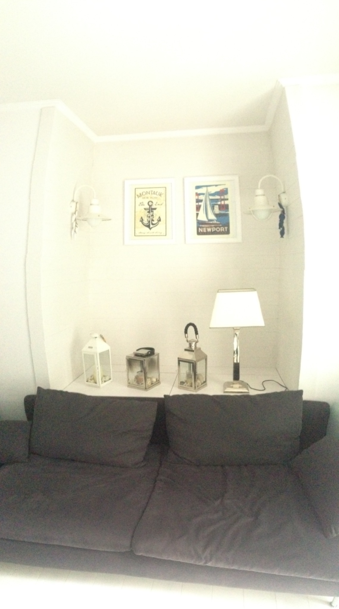 Nadmorski apartament: Szara sofa z poduszkami przed białą półką z lampionami, lampą i marynistycznymi obrazkami w ramkach.