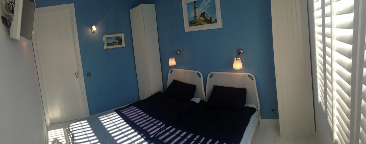 Nadmorska sypialnia z dwoma łóżkami, niebieskie ściany, białe meble i żaluzje przepuszczające promienie słoneczne. Na ścianie obrazek z motywem latarni morskiej.