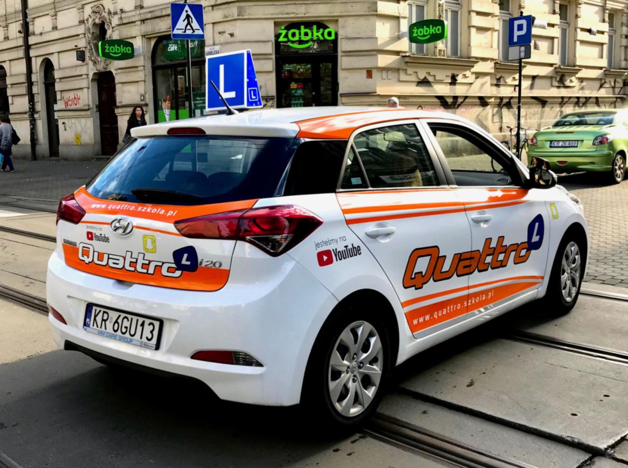 Biały samochód z pomarańczowymi akcentami i logo Quattro L, zaparkowany na ulicy z widocznym znakiem 'L' i szyldami sklepów Żabka w tle.
