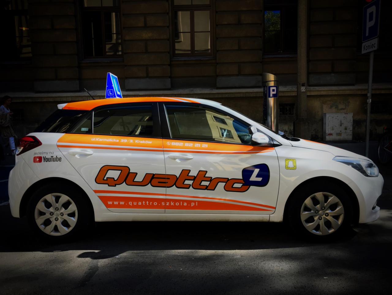 Samochód oznaczony logo i danymi kontaktowymi firmy oferującej kursy nauki jazdy Quattro, zaparkowany przy ulicy Karmelickiej w Krakowie, z widocznym znakiem 'L' na dachu.