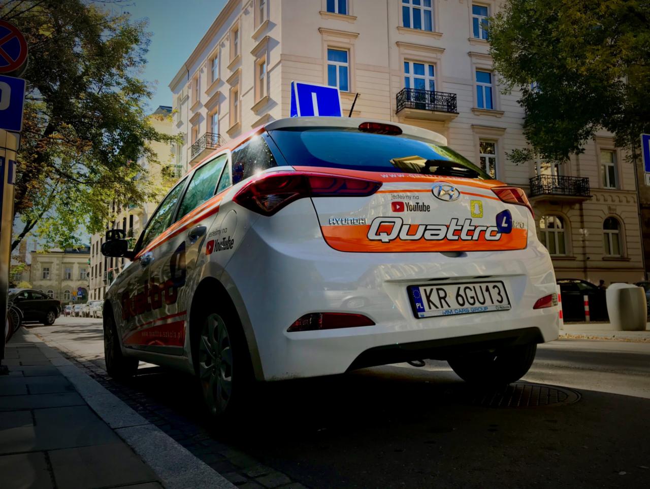 Biały Hyundai i20 z pomarańczowymi pasami i logo szkoły jazdy Quattro L, zaparkowany na chodniku przy ulicy w mieście, widok z tyłu i boku.
