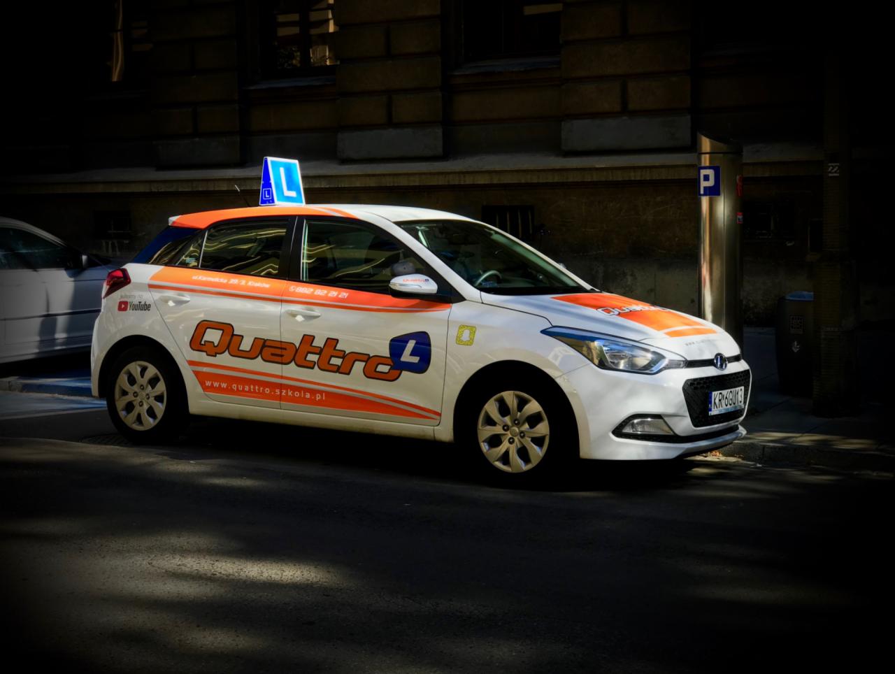 Biały samochód z pomarańczowymi akcentami i literą 'L' na dachu, zaparkowany na ulicy w cieniu budynku, z widocznym logo 'Quattro' i adres strony internetowej na boku.