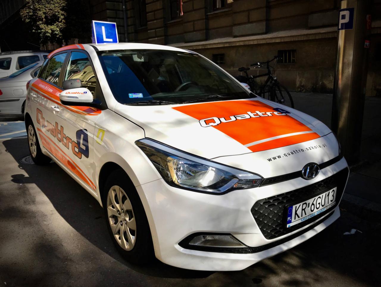 Biały samochód z pomarańczowymi akcentami i znakiem 'L' na dachu, zaparkowany na ulicy miejskiej, z widocznym logo 'Quattro' i adresem strony internetowej.