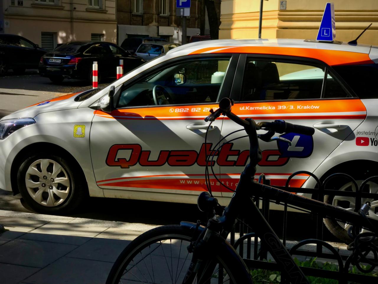 Samochód oznaczony literą 'L' na dachu, zaparkowany przy ulicy, z grafiką 'Quattro' i danymi kontaktowymi, częściowo zasłonięty przez rower.