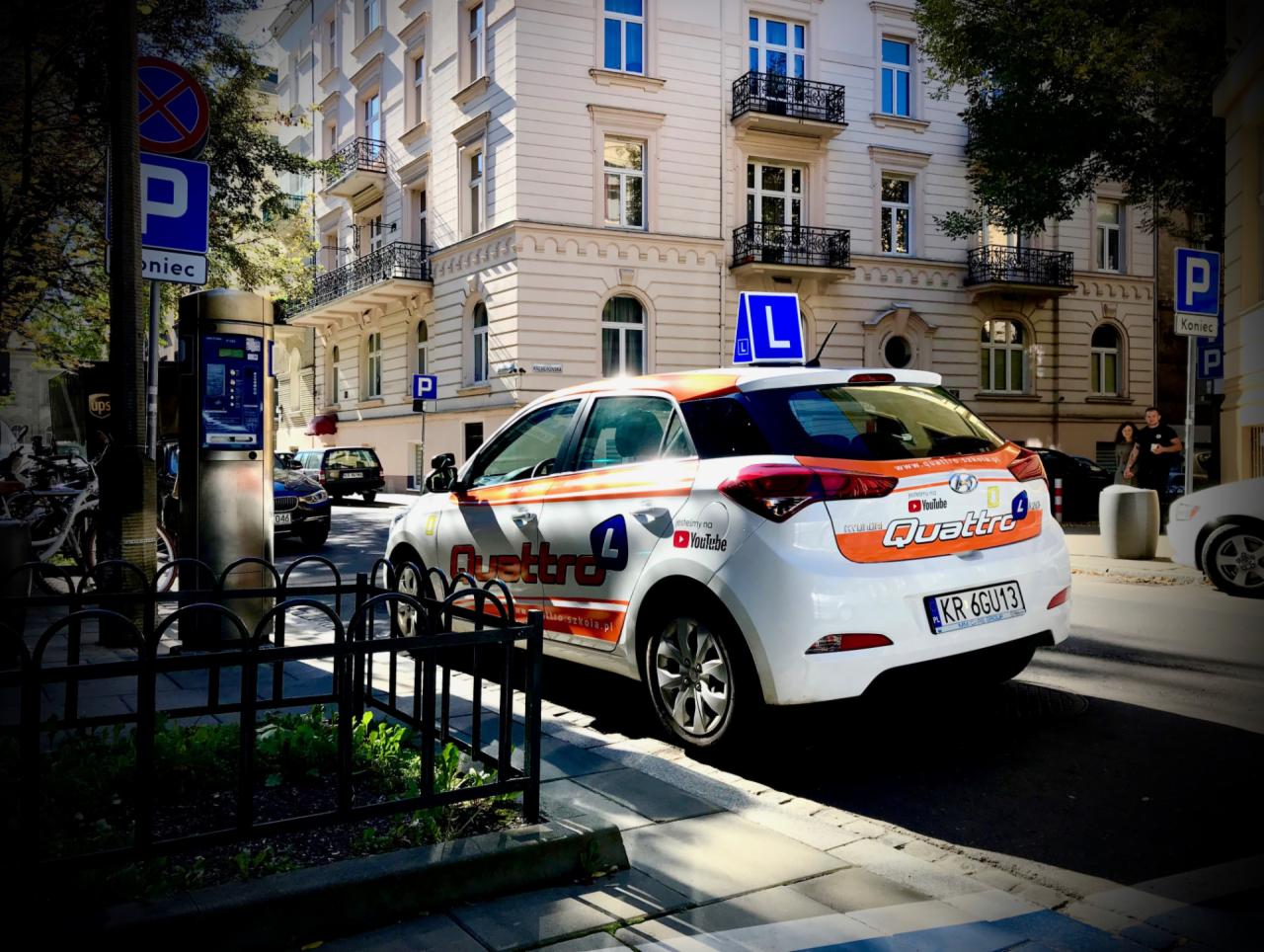 Oznakowany samochód z literą 'L' na dachu, zaparkowany przy ulicy w mieście, z widocznym parkometrem i budynkiem w tle. Auto oklejone reklamami z logo 'Quattro' i odnośnikami do YouTube.