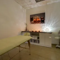 Jasne wnętrze gabinetu z ł&oacute;żkiem do masażu, szafką z ręcznikami i urządzeniem do zabieg&oacute;w na ciało, oświetlone świecami i lampą dekoracyjną.