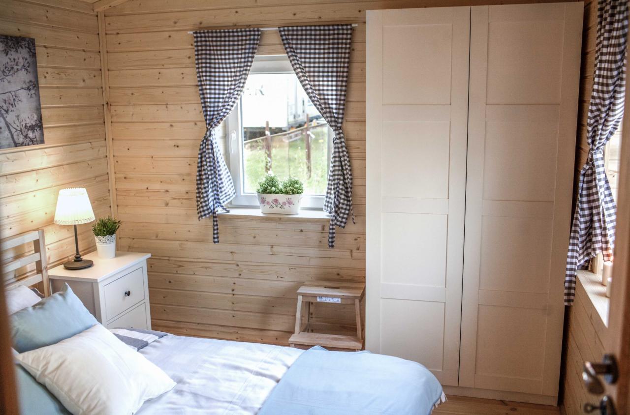 Przytulna sypialnia w domku z drewnianych bali, z łóżkiem przykrytym błękitną narzutą, białymi poduszkami, białą szafką nocną z lampką i rośliną, oknem z zasłonami w kratkę oraz białą szafą.