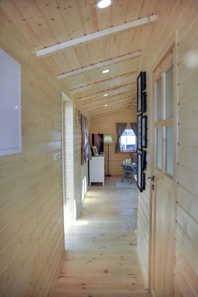 Wnętrze domu wykończonego boazerią, widok z korytarza na salon z telewizorem, lampą podłogową i stolikiem kawowym przy oknie z zasłonami w kratę.