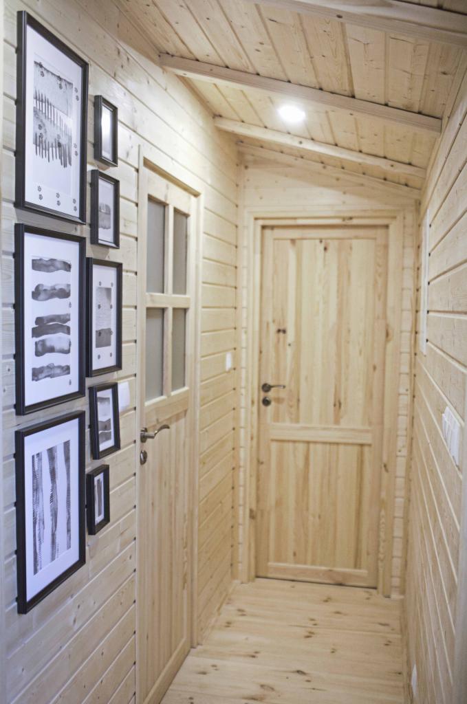Wnętrze domu szkieletowego: korytarz z drewnianą boazerią, galerią czarno-białych grafik w ramkach, drewnianymi drzwiami i podłogą z desek.